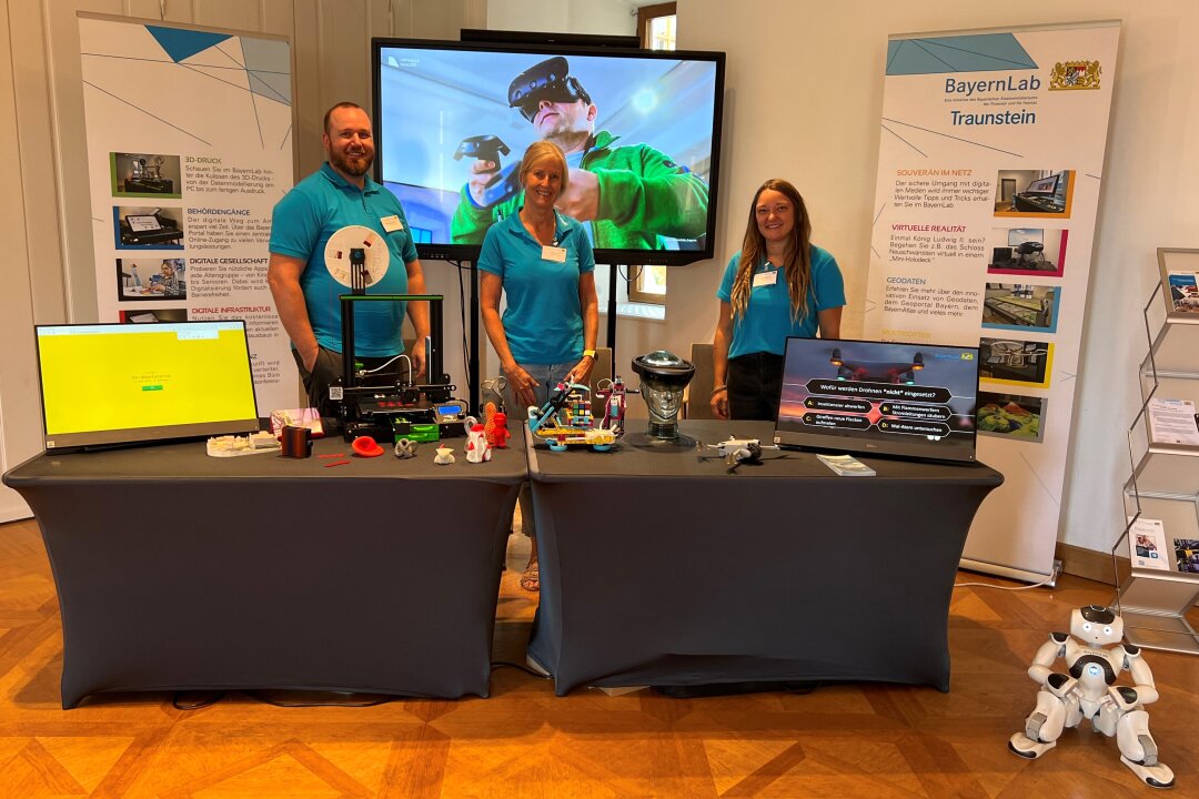 Zwei Frauen und ein Mann vom BayernLab stehen hinter einem Präsentationstisch mit digitalen Exponaten und lächeln in die Kamera. Vorne rechts sitzt ein humanoider Roboter.